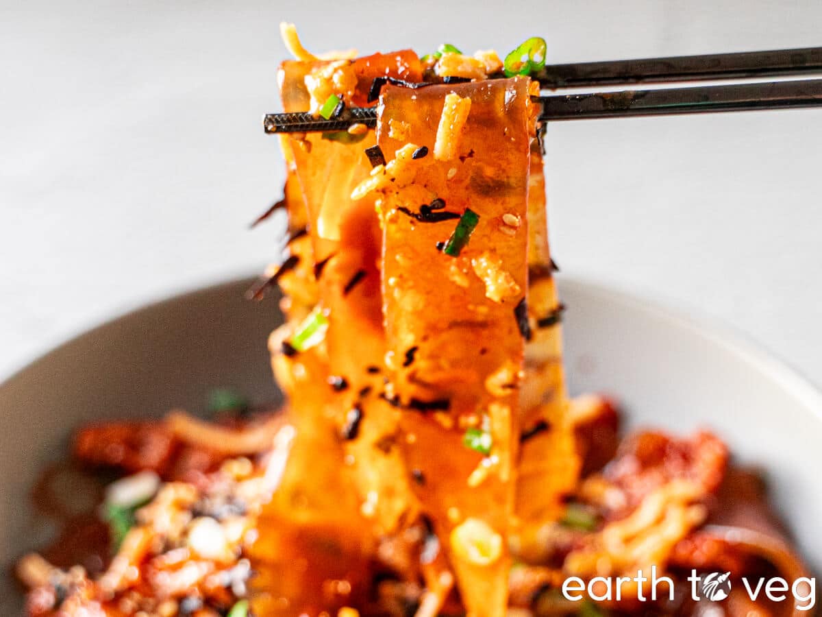 A pair of chopsticks lifts up spicy wide glass noodles from a bowl.